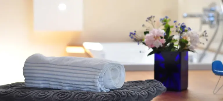 Rolled white towel on a dark textured mat in a spa-like setting, with a blue vase of flowers in the background