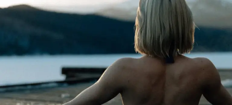 Person with bare shoulders and short hair, seen from behind, sitting by a lake with distant mountains at dusk