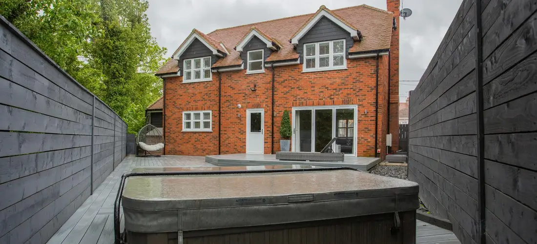 Backyard with a hot tub in front of a red brick house, flanked by tall wooden fences on both sides.