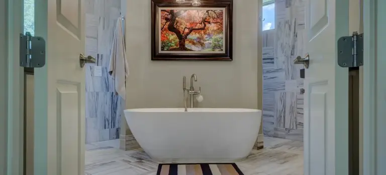 A white freestanding bathtub centered in a marble-tiled bathroom with a framed landscape painting above and double doors open on either side.