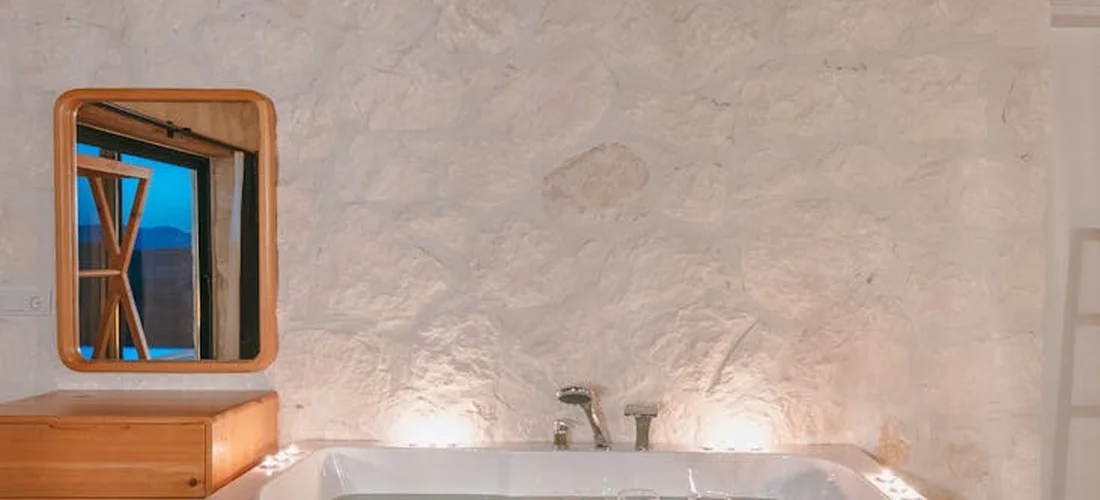 Freestanding bathtub against a textured stone wall with a wooden-framed mirror reflecting a window