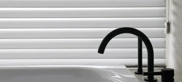 Close-up of a black curved bathroom faucet over a white bathtub with horizontal blinds in the background.