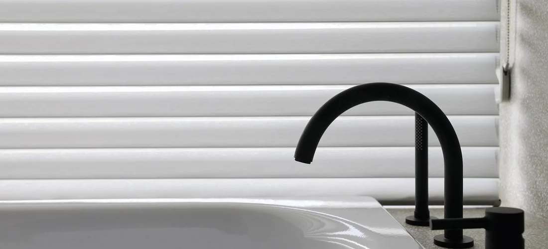 Close-up of a black curved faucet on a white bathroom tub with horizontal blinds in the background