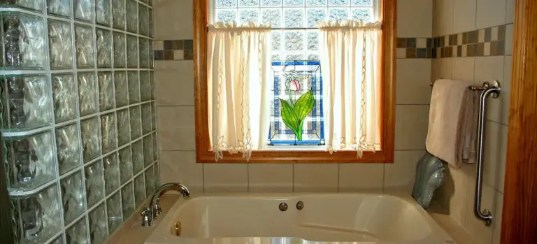 White built-in tub in a tiled bathroom with a glass-block wall on the left, a wooden-framed window with sheer curtains above the tub, and a towel rack on the right.