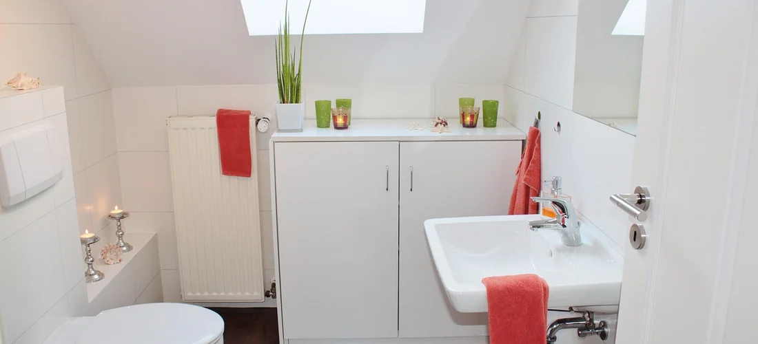 Bright white bathroom featuring a compact vanity, white sink, and red towels; clean, seamless walls suggest PVC or laminate panel materials for shower walls and baths.