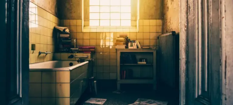 Old tiled bathroom with a bathtub, a small window letting in warm light, and cluttered shelves, conveying a home where energy use matters.