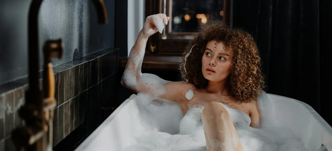 Woman with curly hair sits in a bubble-filled bathtub, looking to the side.