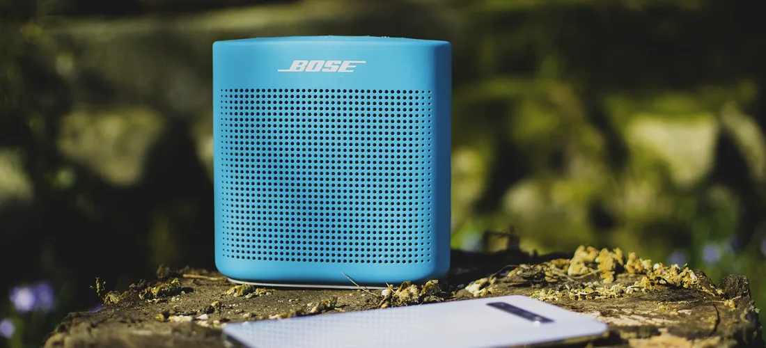 Blue Bose portable speaker on a wooden surface outdoors with a blurred natural background and a notebook nearby.