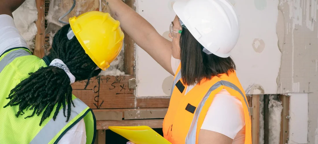 Two construction workers wearing safety vests and hard hats review plans at a partially finished indoor space.