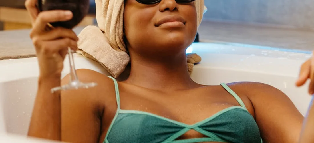Individual relaxing in a bathtub, wearing a teal bikini top, with a towel wrapped around their head, holding a glass.
