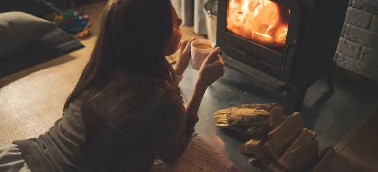 A person sits on the floor by a glowing fireplace, holding a mug, with stacked firewood nearby in a cozy living room.