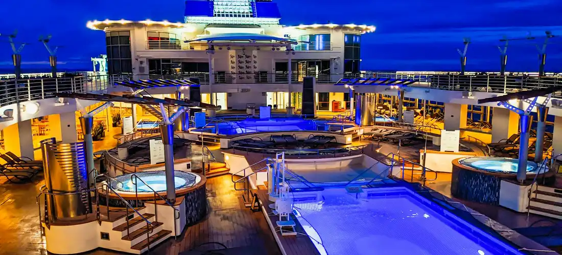 Nighttime view of a cruise ship pool deck with several hot tubs and a central pool, illuminated with blue lights.