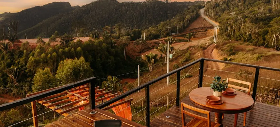 Outdoor wooden deck with a round table and chairs, overlooking rolling hills and trees; a railing frames a scenic valley.