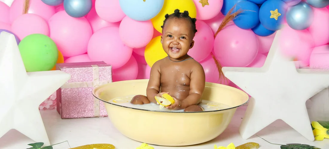 A baby sitting in a yellow tub at a party, surrounded by pink and blue balloons, star-shaped decorations, and gift boxes.