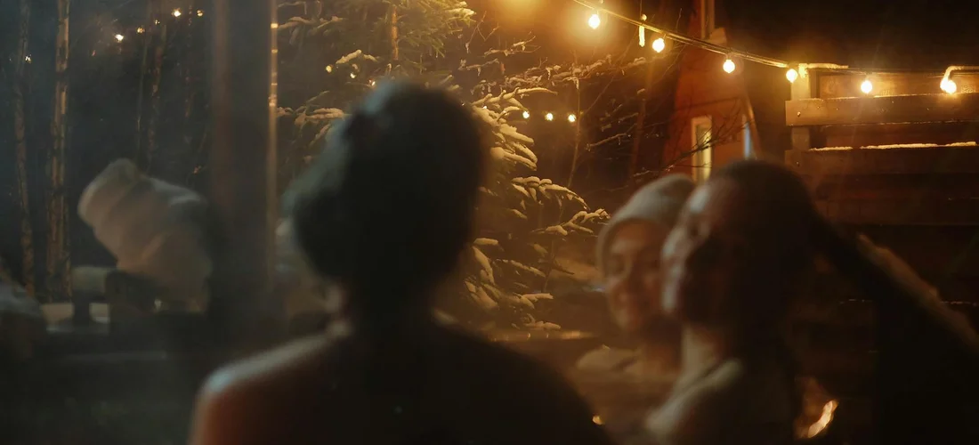 A group of people gathered around a warm fire pit at night under string lights, creating a cozy outdoor scene.