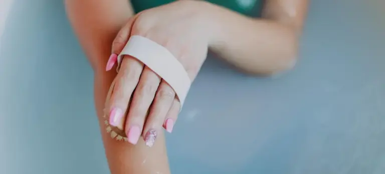 Close-up of a hand with pink nail polish and a white wrist band resting near a blue-tinted bath.