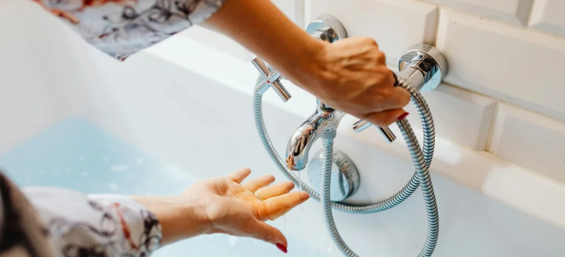 Close-up of hands repairing a bathroom faucet with a flexible shower hose visible in the background.