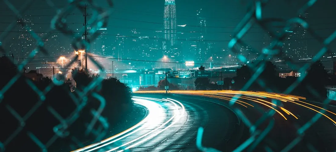 Nighttime city skyline visible through a chain-link fence, with light trails from moving cars.