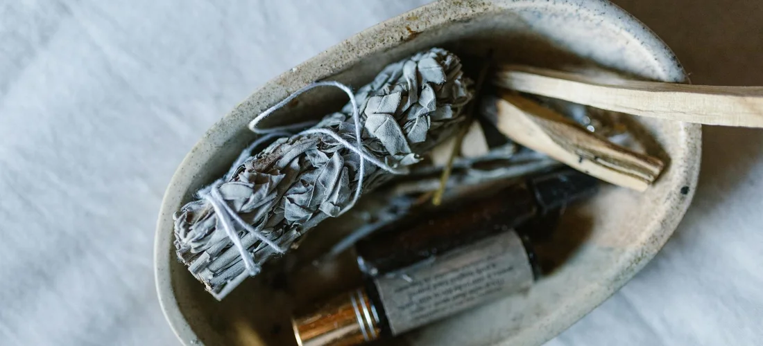 Close-up of a metal container with a bundle of dried herbs tied with string, an amber essential oil bottle, and a wooden stir stick, representing hot-tub aromatherapy products.