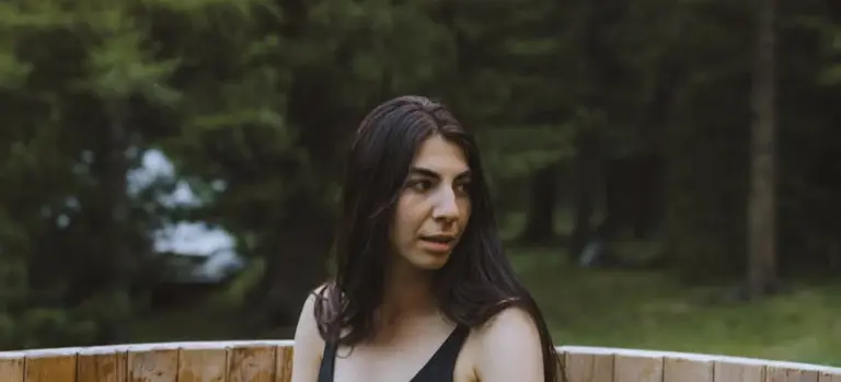Woman with long dark hair standing outdoors near a wooden hot tub, with trees in the background.