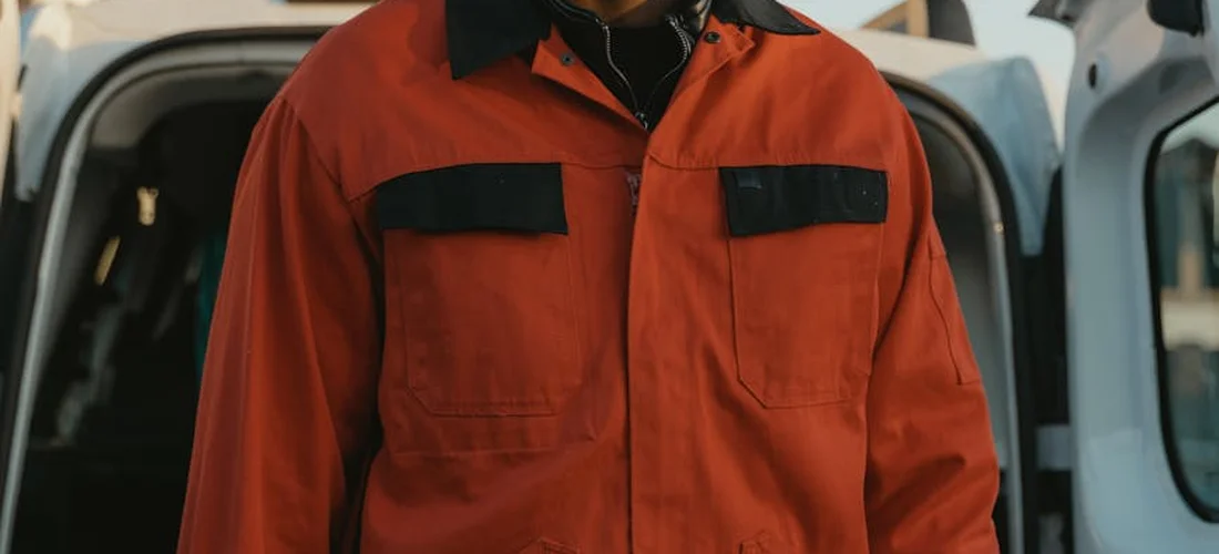 Worker in bright orange protective coveralls standing near an open vehicle, illustrating PPE for maintenance and electrical safety during wet work.