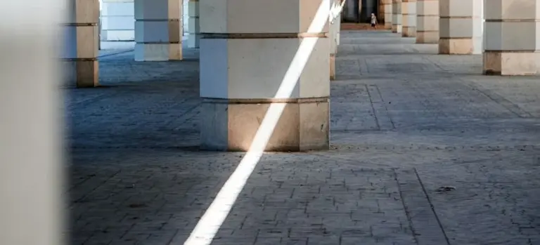Interior of a columned concrete structure with a bright diagonal beam of light crossing a textured paved floor.