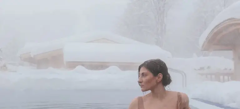 A woman relaxing in an outdoor hot tub in a snowy setting, with snow-covered buildings in the background.