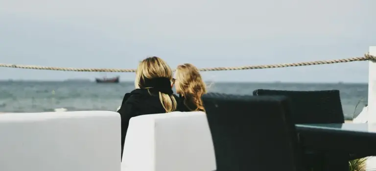 Two people sitting on a modern outdoor deck facing the water, with white seating and dark chairs.