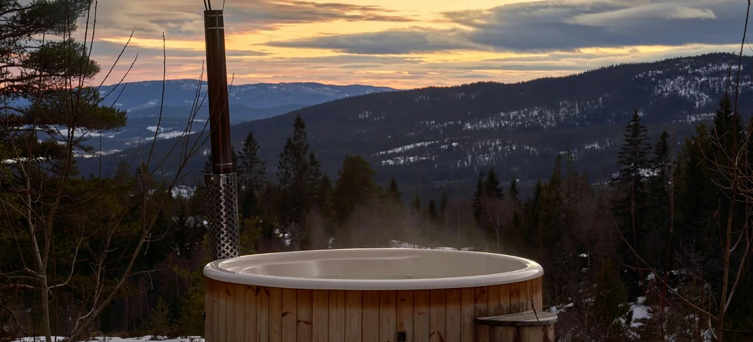 Wooden hot tub on a deck in a snowy mountain landscape at dusk, with pine trees and distant hills.
