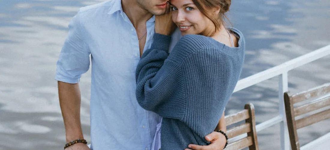 A couple stands on a waterfront deck near a white railing, embracing each other with wooden benches nearby.