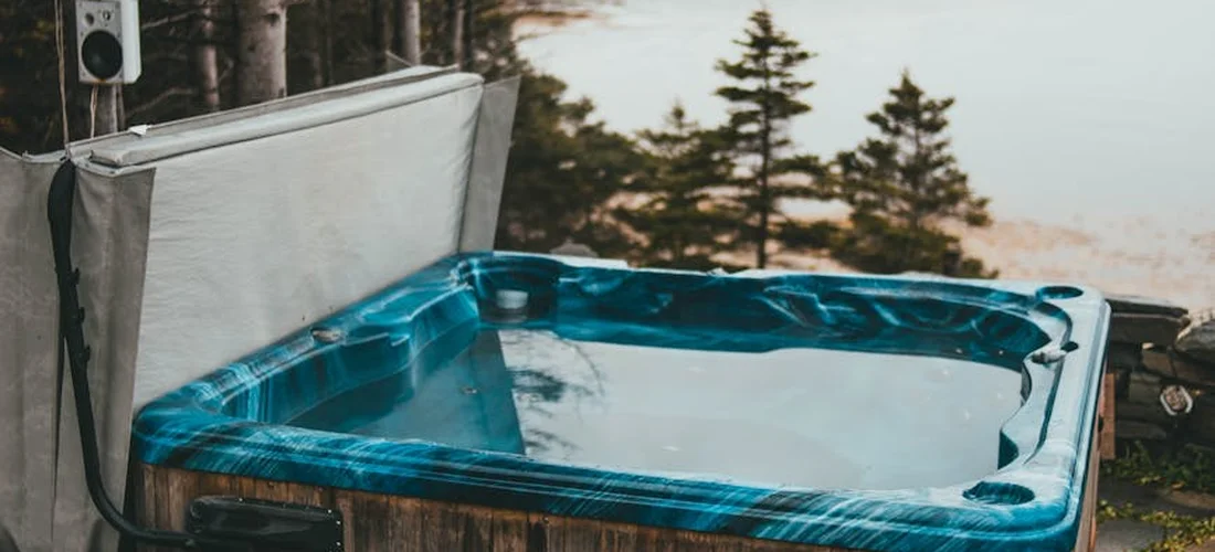 Outdoor hot tub with blue water and an open lid, set among trees by a lake