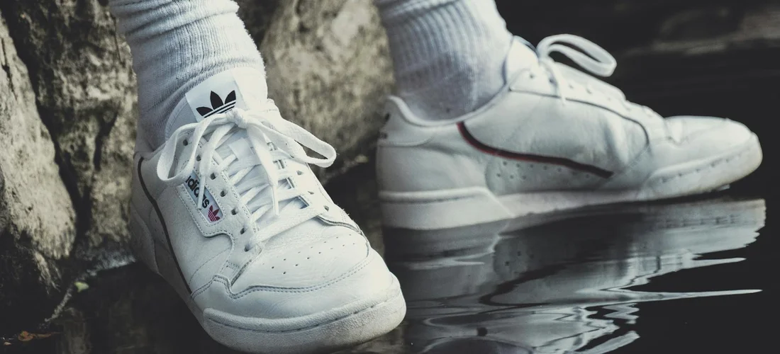 White athletic sneakers with socks resting on a wet, reflective surface beside a rock wall