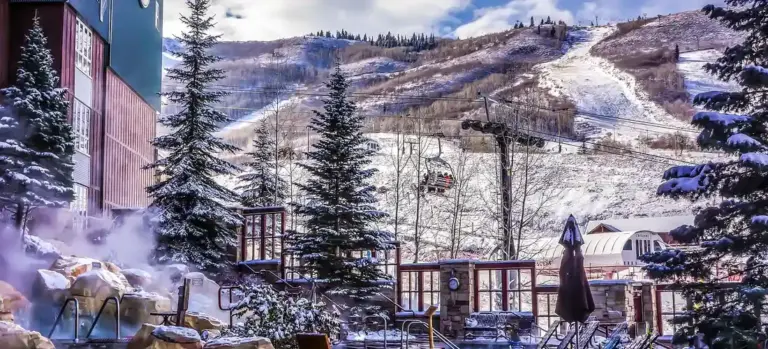 Outdoor hot tub setup in a snowy mountain resort with steam rising from the water, evergreen trees, and snow-covered slopes in the background.