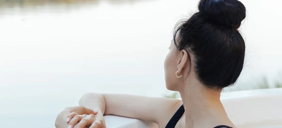 Person with dark hair tied in a bun resting on the edge of a pool or hot tub, viewed from behind.