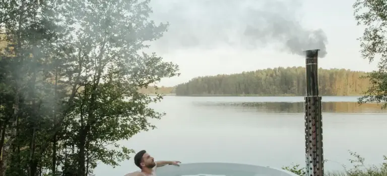 Person relaxing in a hot tub by a lake with trees in the background; steam rising from the tub.