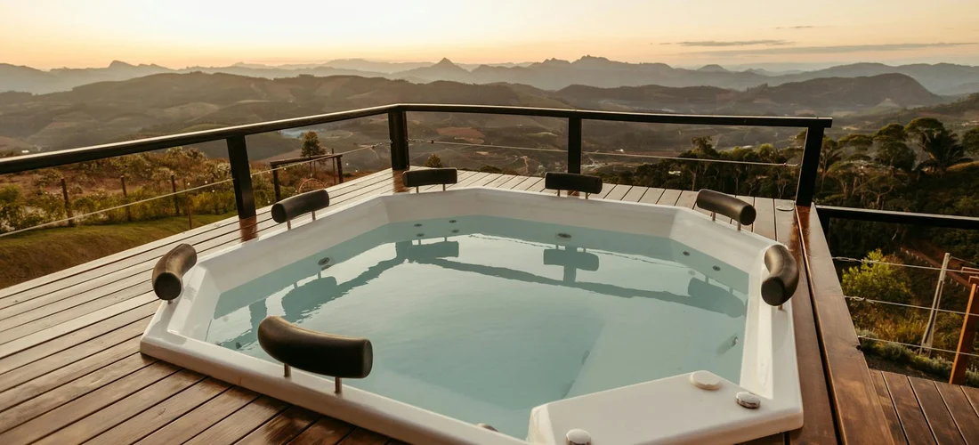 A white hot tub on a wooden deck with glass railing, set against a mountainous landscape at sunset.