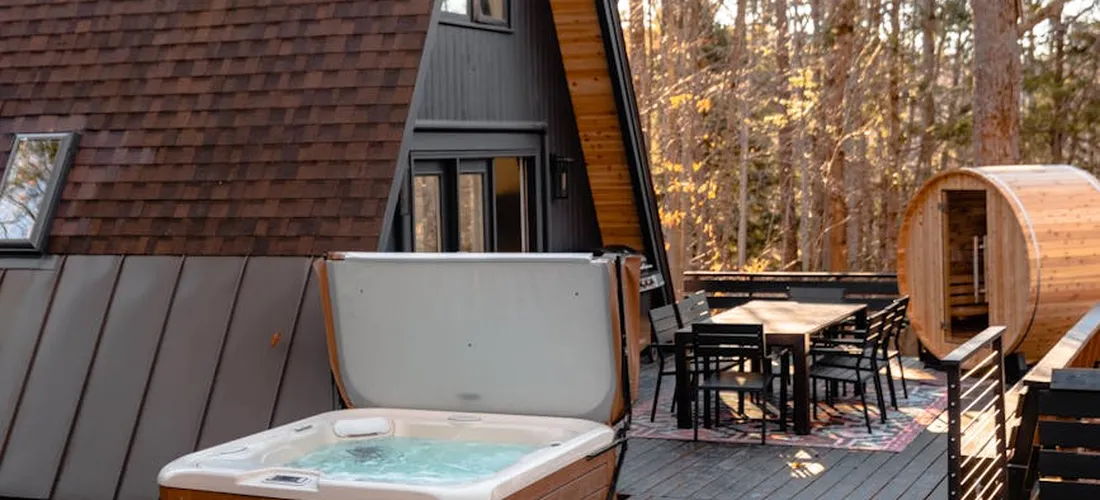 Outdoor hot tub on a wooden deck with patio furniture and a wooden sauna in the background, surrounded by trees.