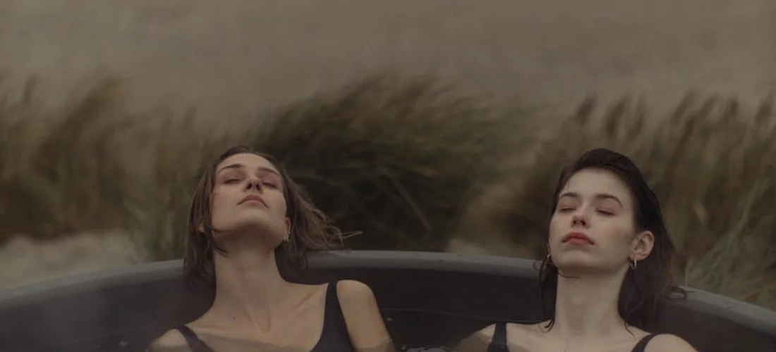 Two people sit in a hot tub outdoors with tall grasses in the background.