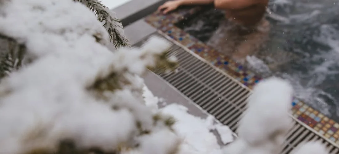 Outdoor hot tub in winter with snow on nearby branches in the foreground and a drain grate visible along the tub edge.