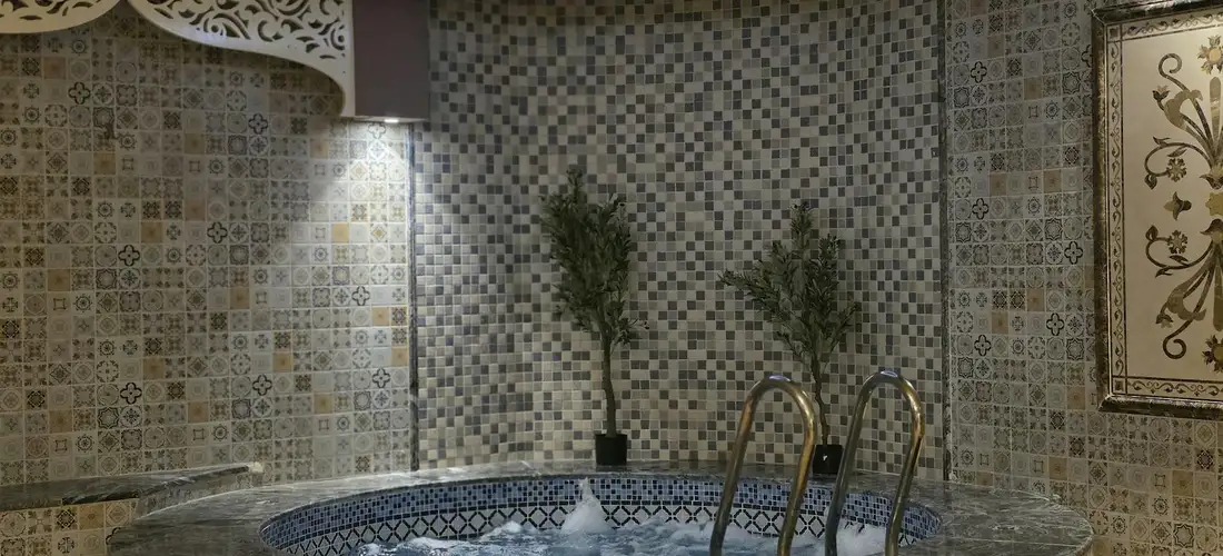 Blue mosaic-tiled hot tub in a bathroom, with metal handrails and bubbling water.