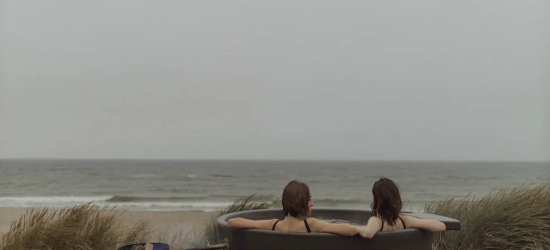 Two people sit in an outdoor hot tub by the beach on a cloudy day.