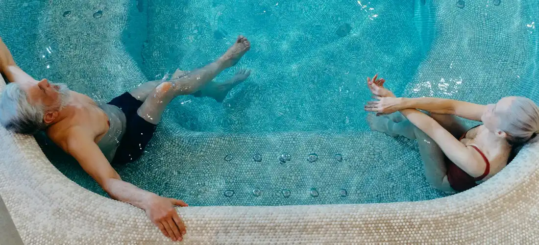 Two seniors relaxing in a hot tub, one on the left leaning back and the other on the right resting their arms on the edge.