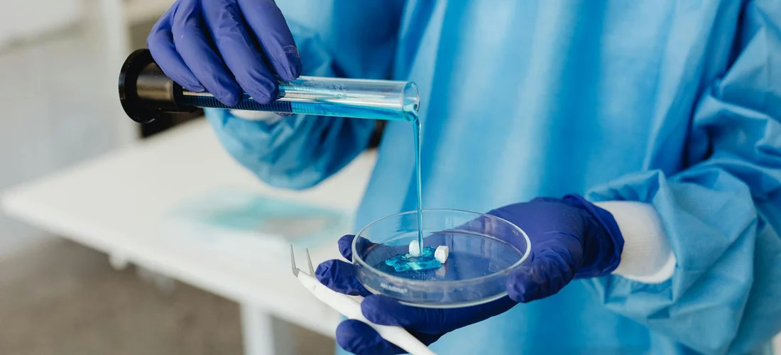 Lab technician wearing blue gloves and a blue lab coat uses a pipette to transfer liquid into a small petri dish.