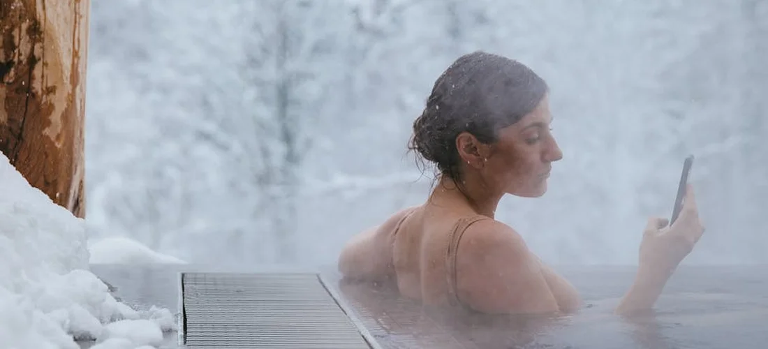 Person soaking in an outdoor hot tub in winter snow, steam rising, holding a smartphone.