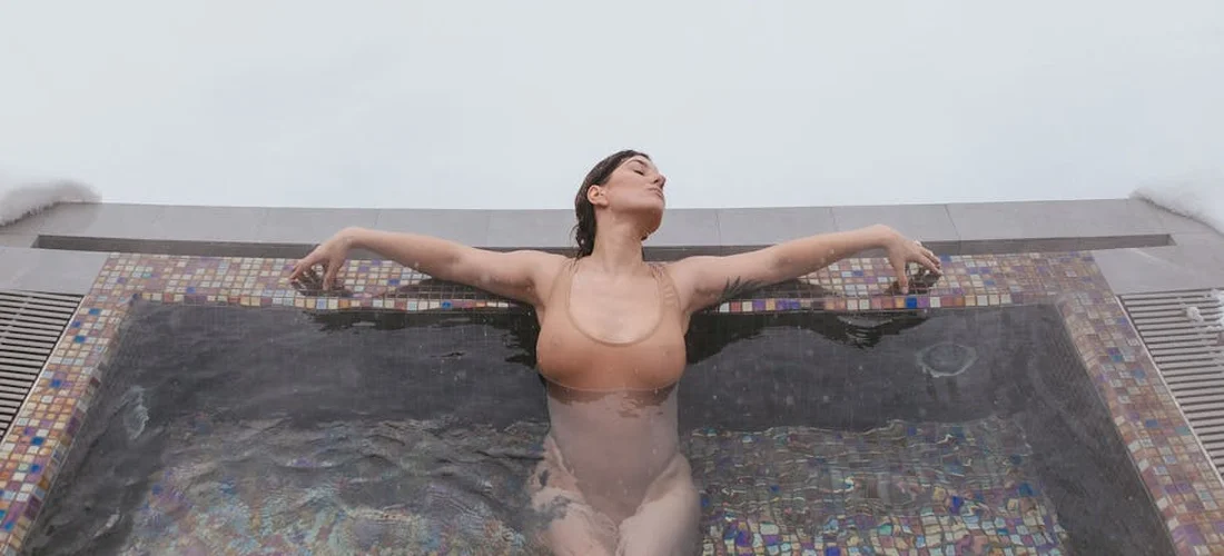 Woman in a brown swimsuit leaning on the edge of an outdoor hot tub, with snow around and looking upward.