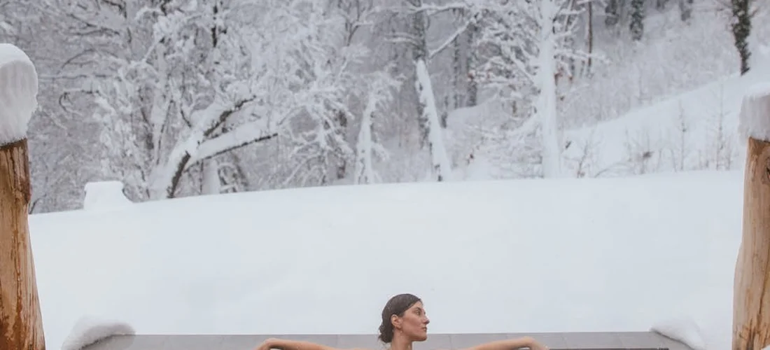 A person relaxing in an outdoor hot tub surrounded by snow-covered trees, with wooden posts framing the scene.