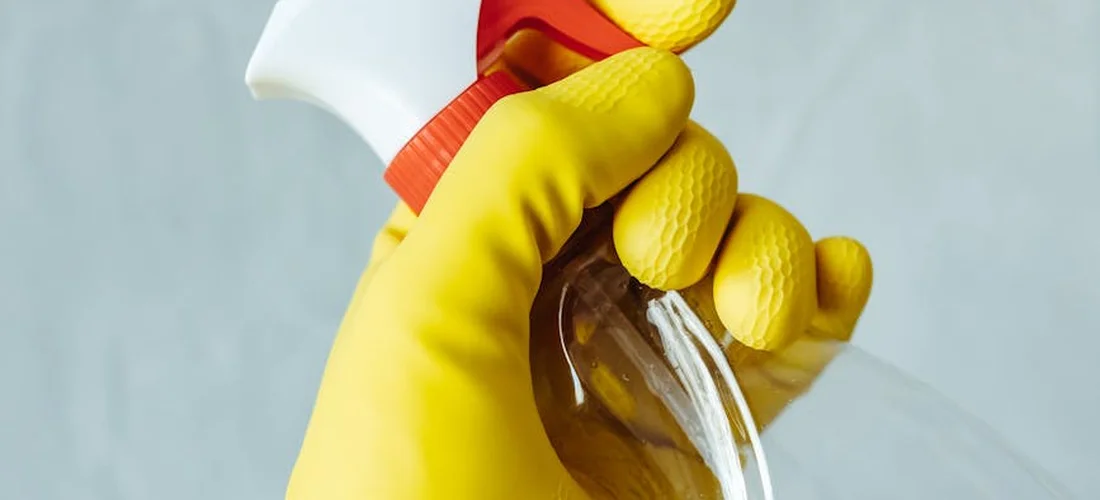 Close-up of a yellow-gloved hand gripping a spray bottle, preparing to spray a cleaning solution.