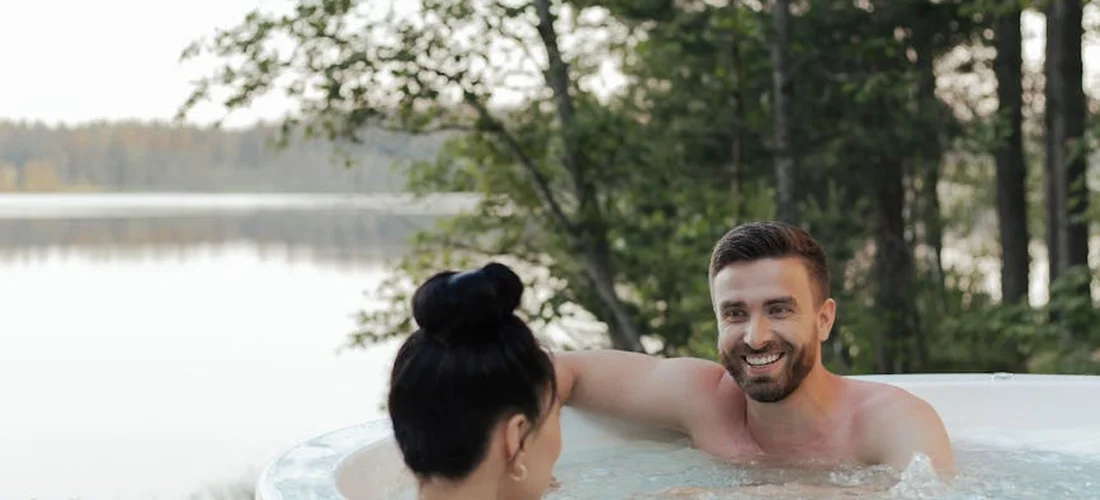 Two adults are relaxing in an outdoor hot tub, smiling at each other with trees and water in the background.