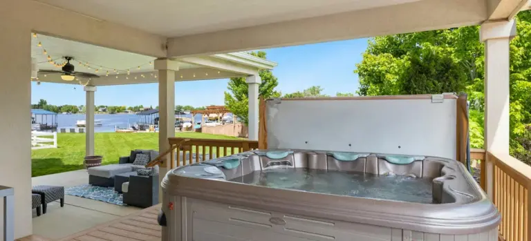 Outdoor backyard with a large hot tub on a wooden deck under a covered patio, string lights, seating area, and a lake in the distance.