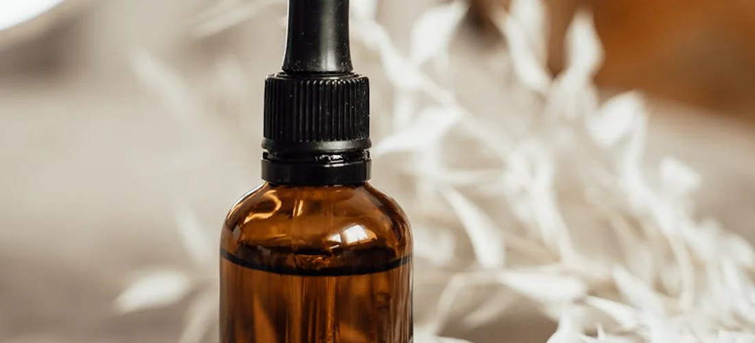 Amber glass dropper bottle with a black cap in sharp focus, with soft white towels blurred in the background.